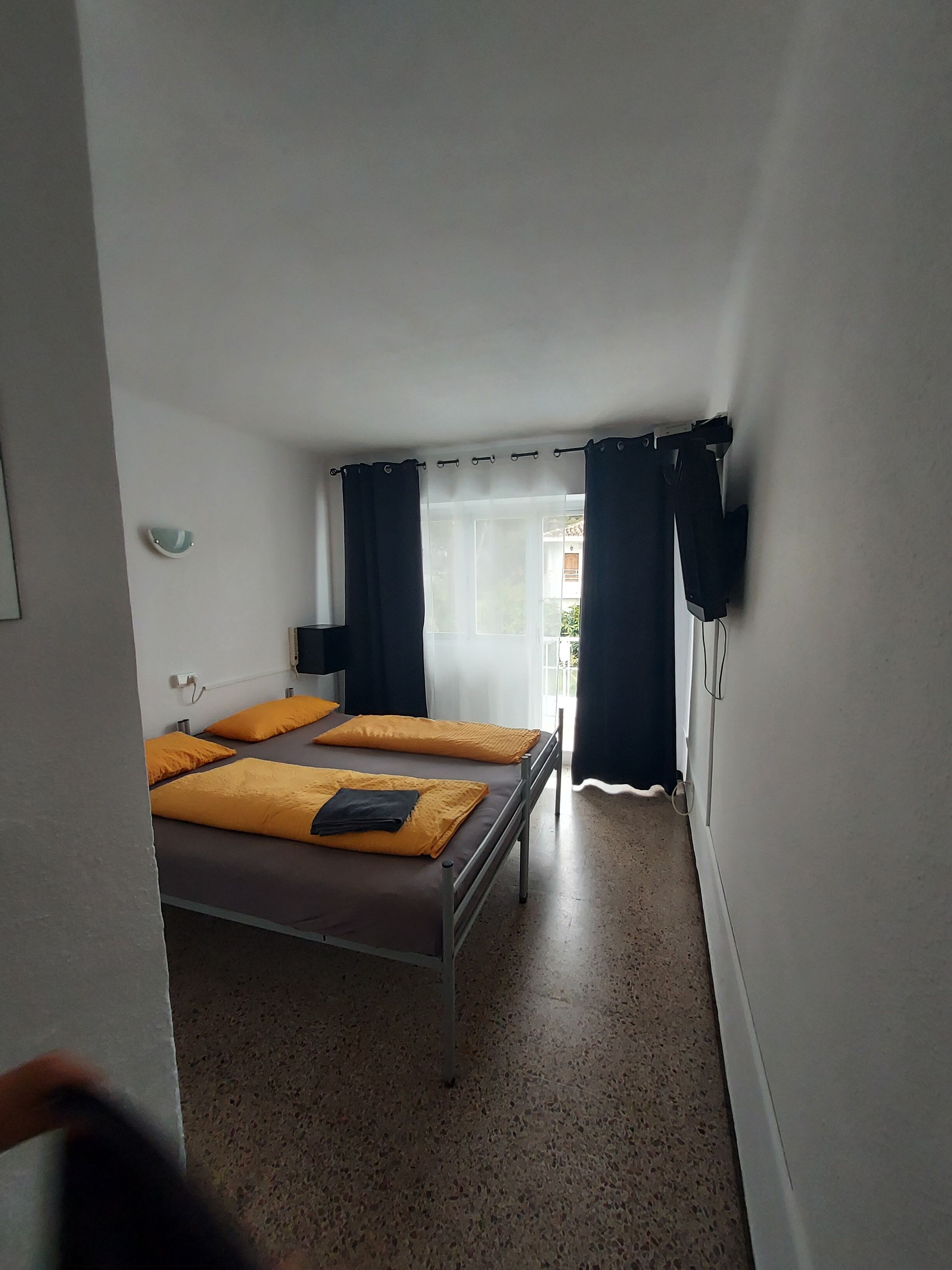 Quarto com cama, mesa de cabeceira, janela com cortinas e vista para o exterior.