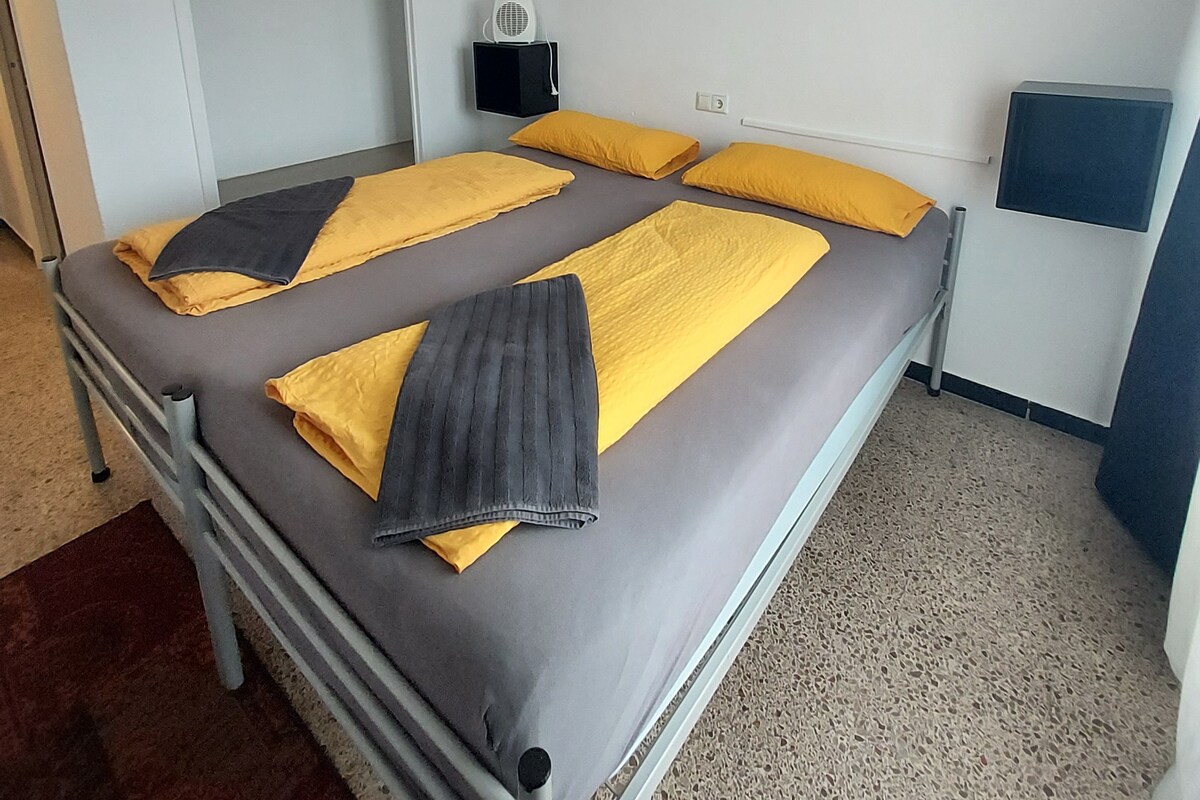 A single bed with a grey and yellow bedspread, a small bedside table with a lamp, and a wall-mounted speaker.