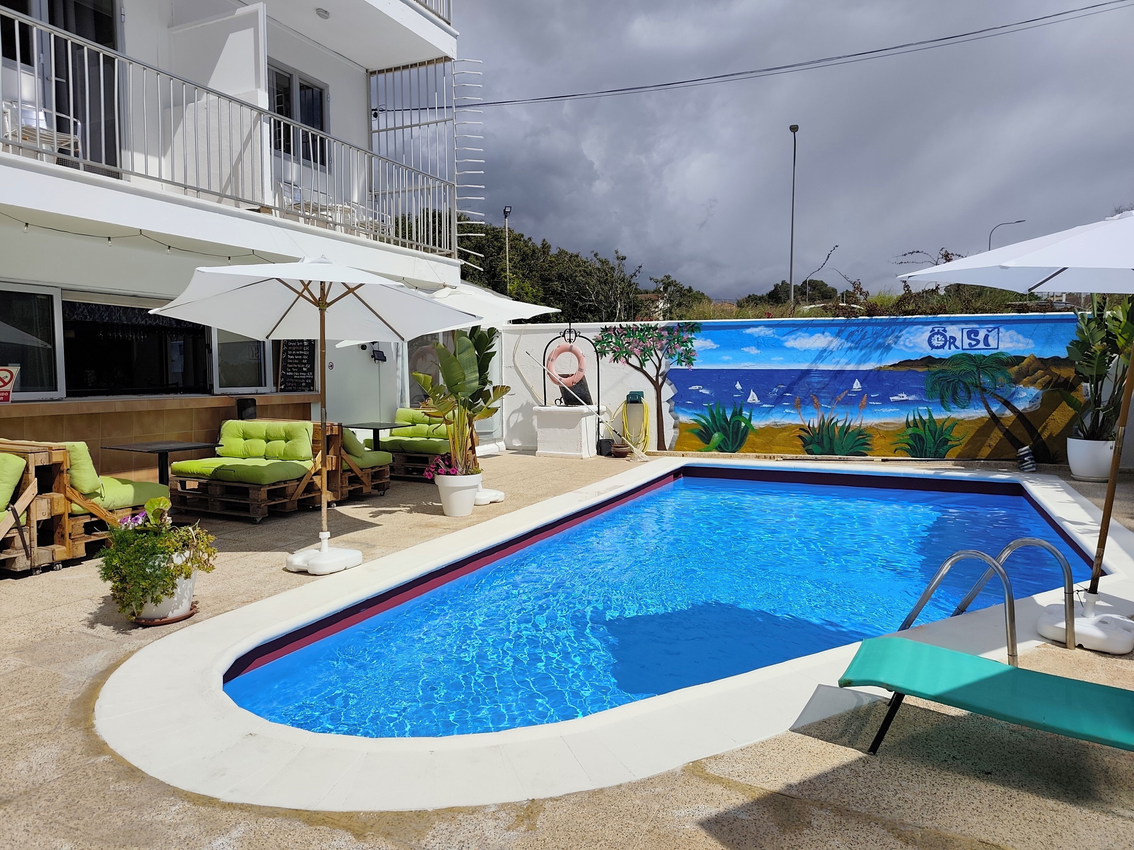 Una piscina con un mural, sillas reclinables verdes y un edificio blanco con balcón.