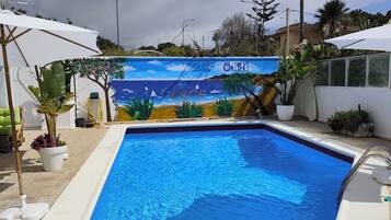 Una piscina con un mural en la pared, rodeada de macetas con plantas y sombrillas.