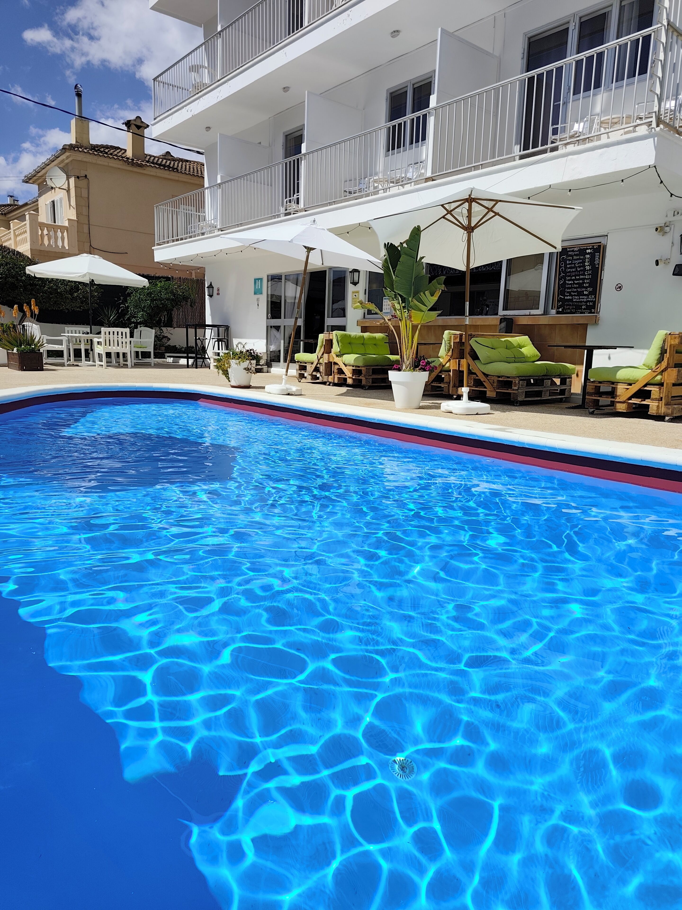 A swimming pool with clear blue water, surrounded by lounge chairs and umbrellas, and a building with balconies and outdoor seating.