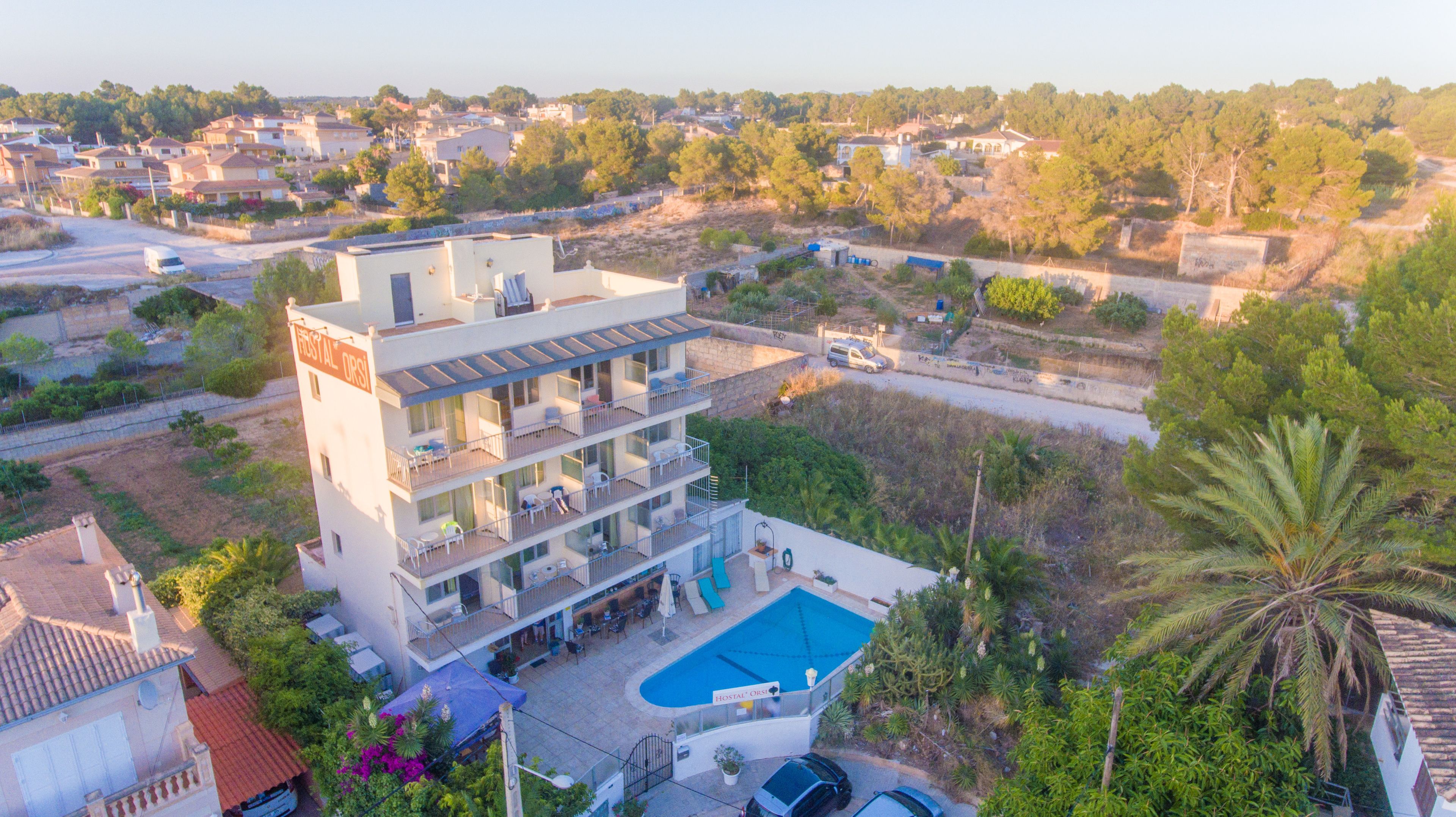 Un hotel de varios pisos con piscina, rodeado de vegetación y edificios residenciales.