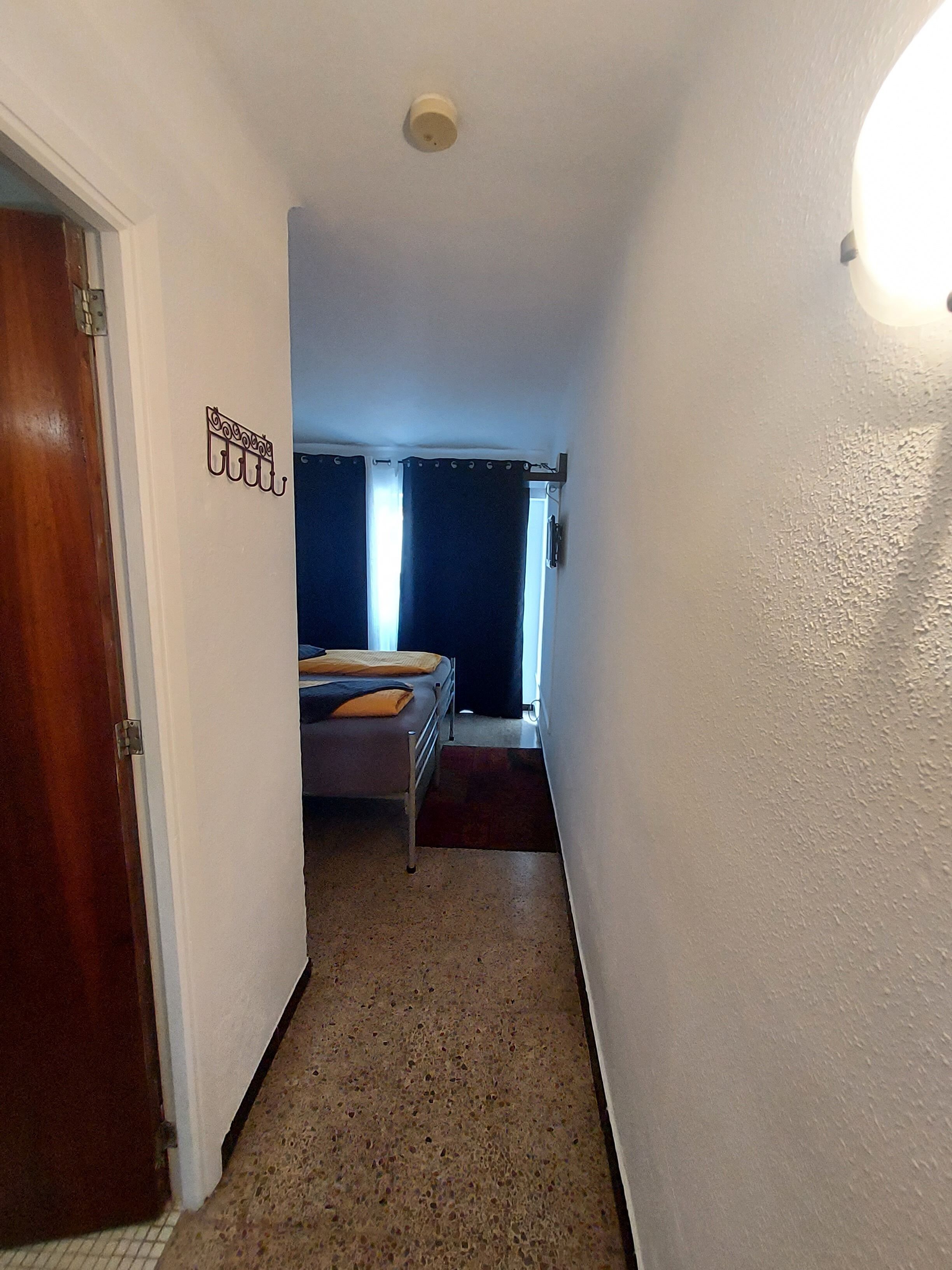 A narrow hallway with a bed in the background, a door on the left, and a light fixture on the right.