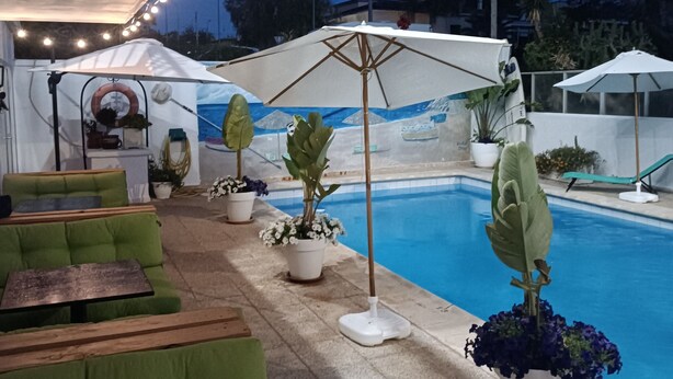 Zona de piscina al aire libre con asientos acolchados de color verde, sombrillas y luces colgantes.