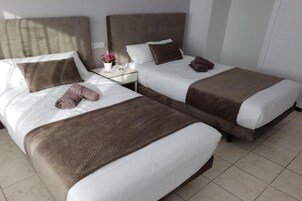 Habitación de hotel con dos camas, cada una con cabecero marrón, ropa de cama blanca y una manta marrón. Hay una mesita de noche con una lámpara y un florero con flores.