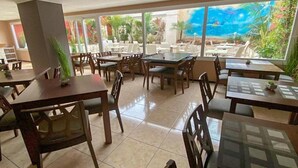 A restaurant with multiple tables and chairs, large windows with a view of palm trees and a mural, and a tiled floor. - Xapala (MALLORCA)