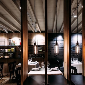 Interior de restaurante con vigas de madera, lámparas colgantes y una zona de bar con estanterías.