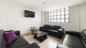 A modern living room with black leather sofas, a glass coffee table, a flat-screen TV mounted on the wall, and a large window providing natural light. - Hotel Dorado 100 (Bogotá)