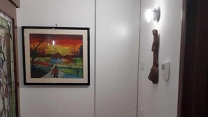 Um corredor com um quadro pendurado na parede, representando uma paisagem de pôr do sol, uma porta branca e uma luminária fixada na parede.