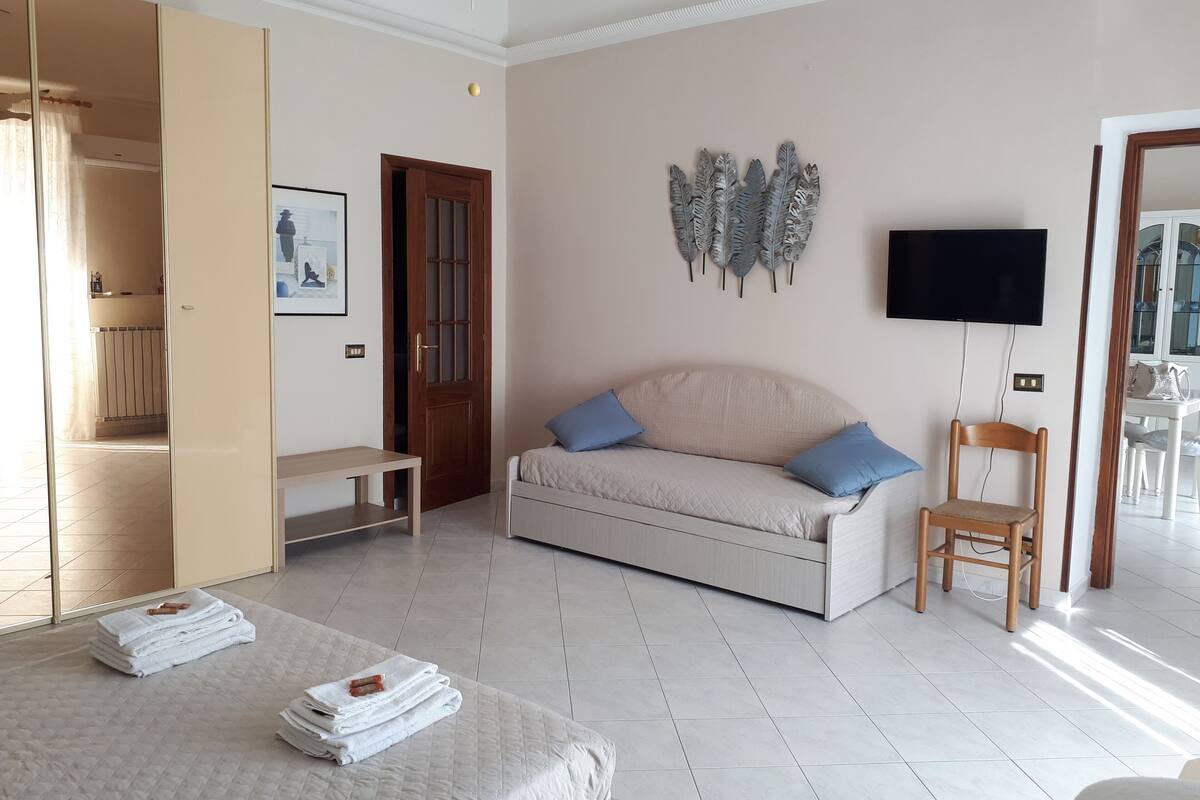 A modern hotel room with a sofa, a wooden chair, a TV, and a bed with towels.