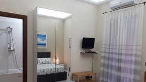 Quarto com cama, guarda-roupa, televisão em uma prateleira, chuveiro e janela com cortinas.