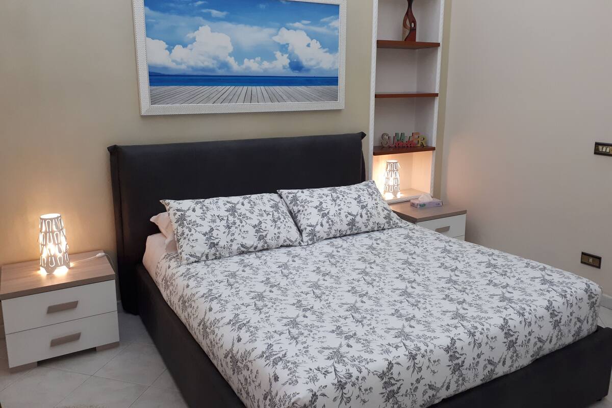A bedroom with a bed, nightstands, a wall-mounted shelf, and a painting of a beach scene.