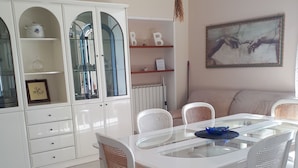 Uma sala de jantar com mesa e cadeiras brancas, um armário com portas de vidro e um quadro na parede.