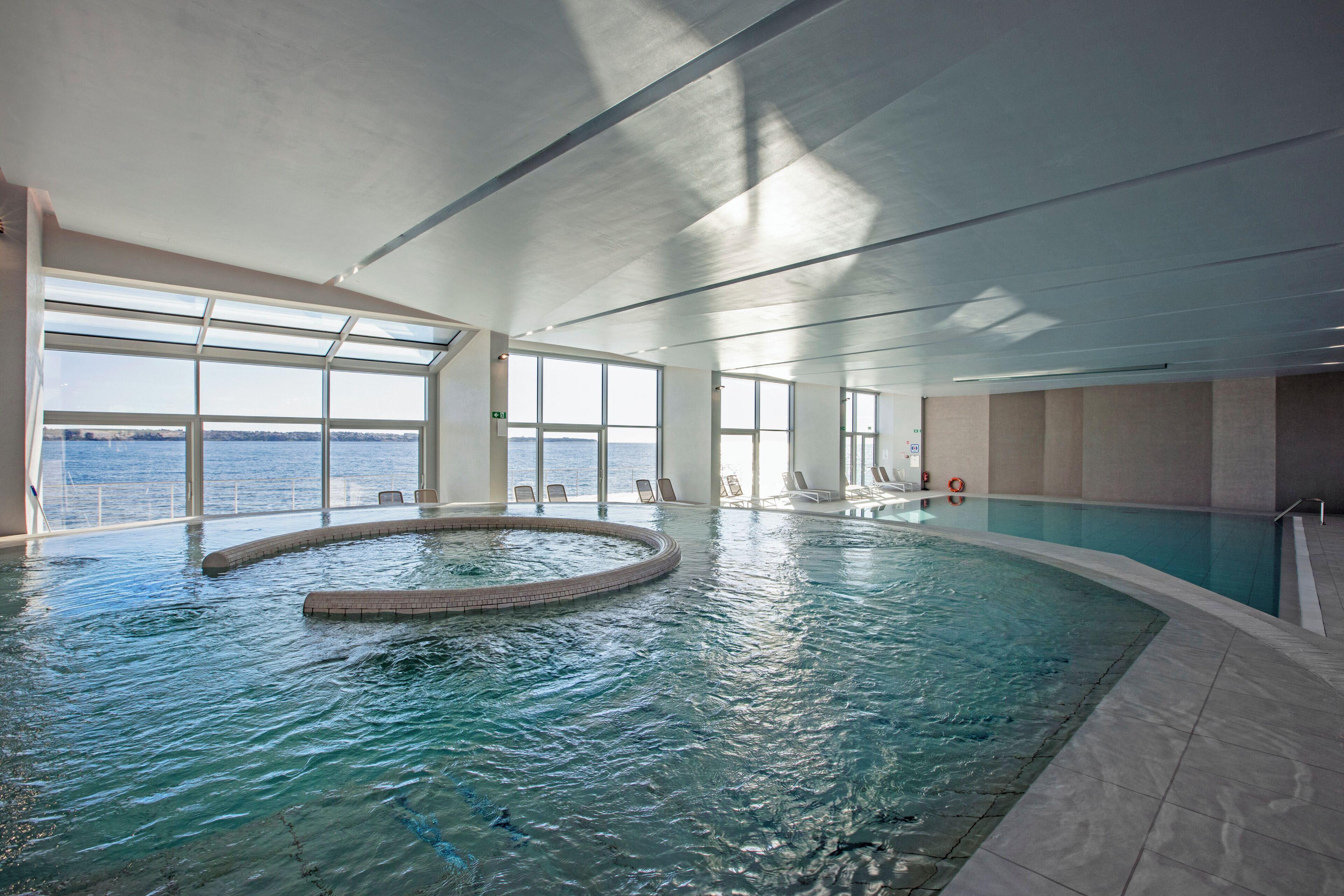 Una piscina interior con vistas despejadas al mar a través de amplios ventanales, un jacuzzi circular y una zona de piscina con sillones.