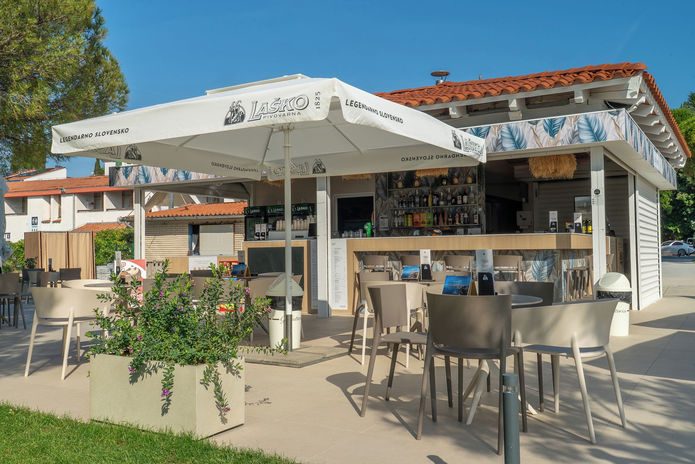 An outdoor seating area with tables and chairs, a bar with a white umbrella, and a building with a red-tiled roof.