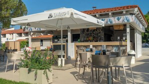 An outdoor seating area with tables and chairs, a bar with a white umbrella, and a building with a red-tiled roof. - Hotel Vile Park Premium (Piran)