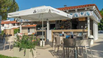 An outdoor seating area with tables and chairs, a bar with a white umbrella, and a building with a red-tiled roof.