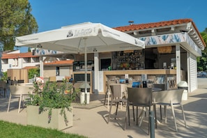An outdoor seating area with tables and chairs, a bar with a white umbrella, and a building with a red-tiled roof. - Hotel Vile Park Premium (Piran)