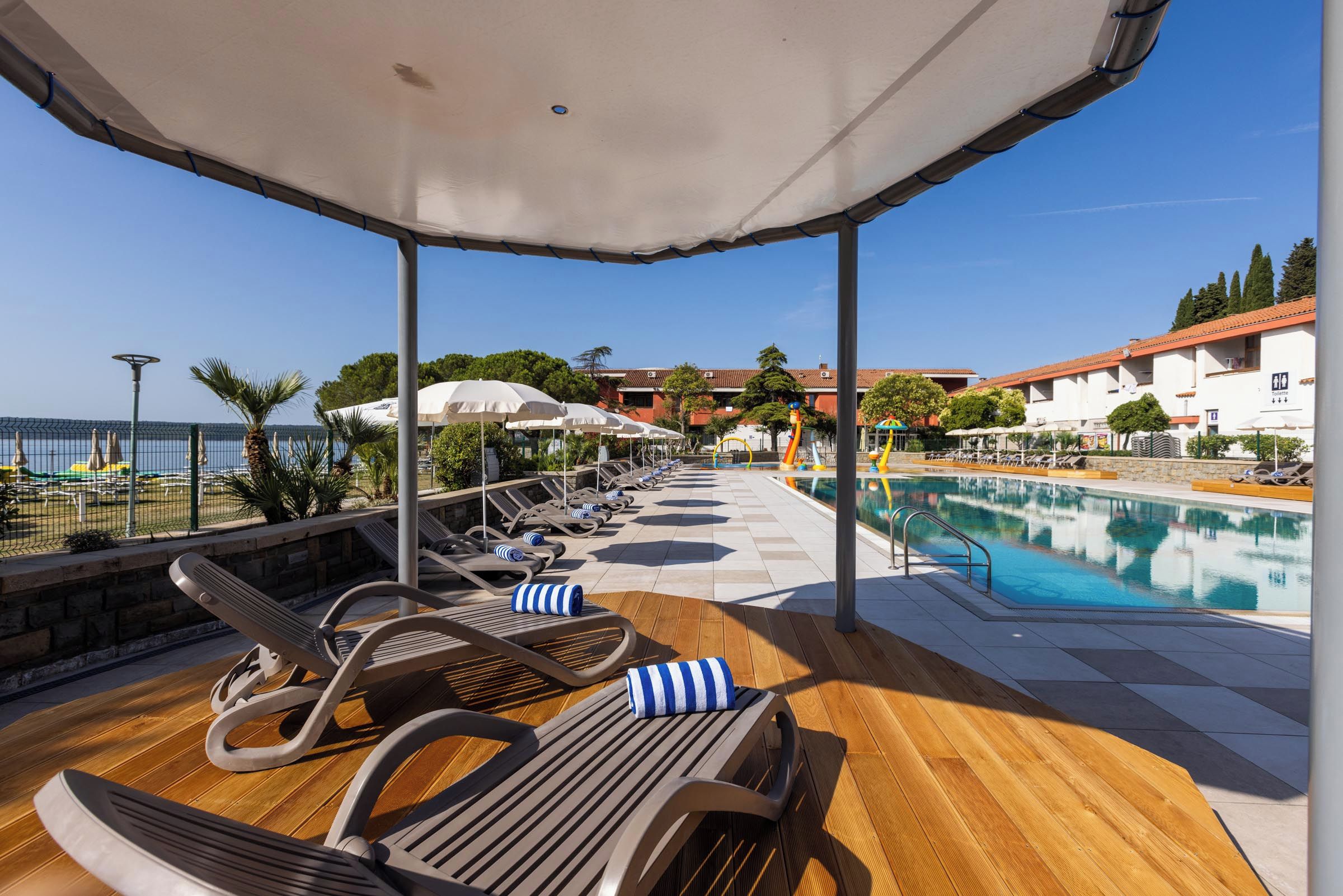 A poolside area with lounge chairs and umbrellas, a wooden deck, and a view of the pool and buildings in the background.