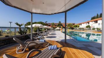A poolside area with lounge chairs and umbrellas, a wooden deck, and a view of the pool and buildings in the background.