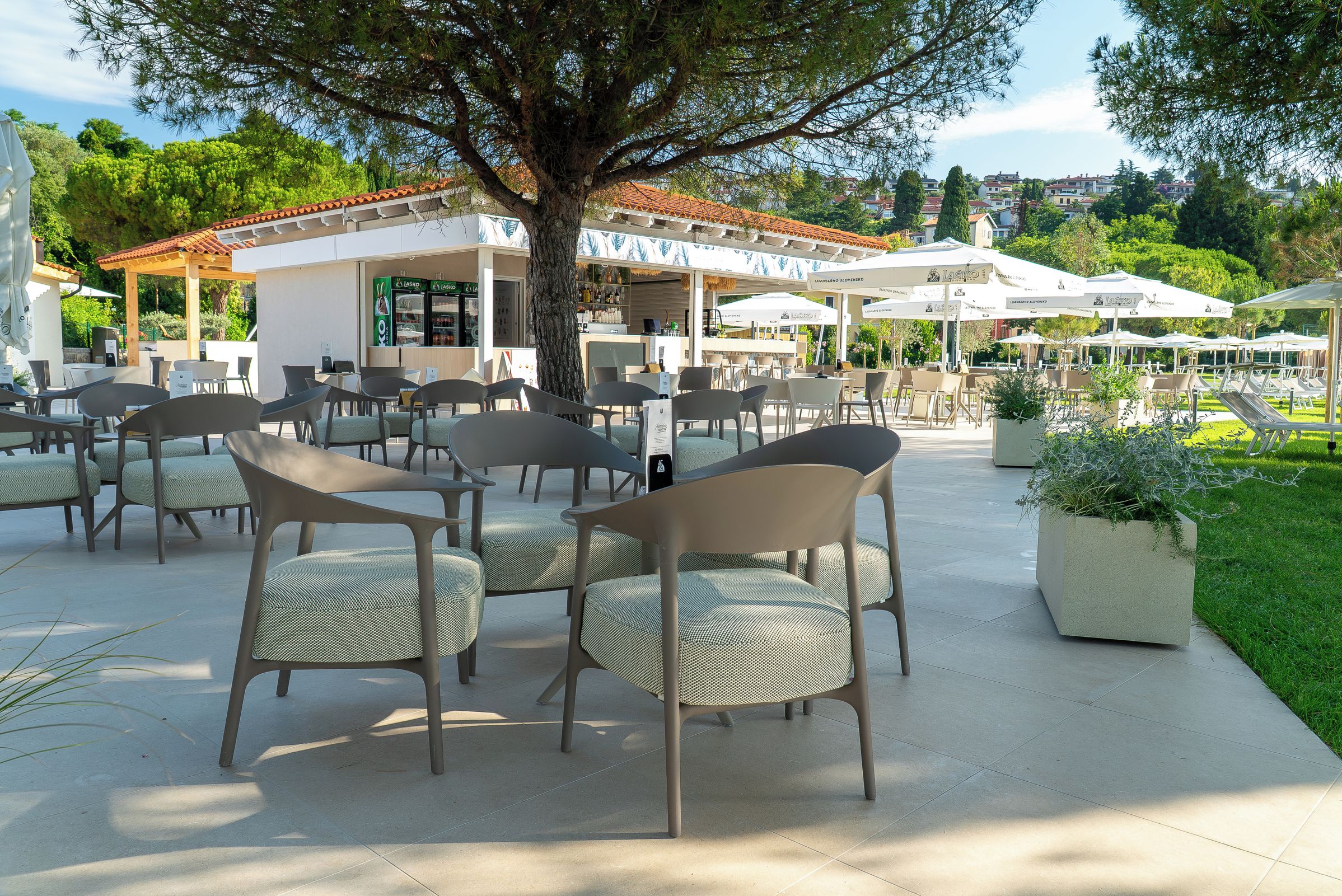 An outdoor seating area with chairs and tables, a building with a tiled roof, and a tree providing shade.