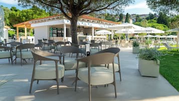 An outdoor seating area with chairs and tables, a building with a tiled roof, and a tree providing shade.