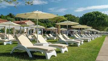 A row of white lounge chairs with beige cushions and large umbrellas, arranged on a grassy lawn with a clear sky above.