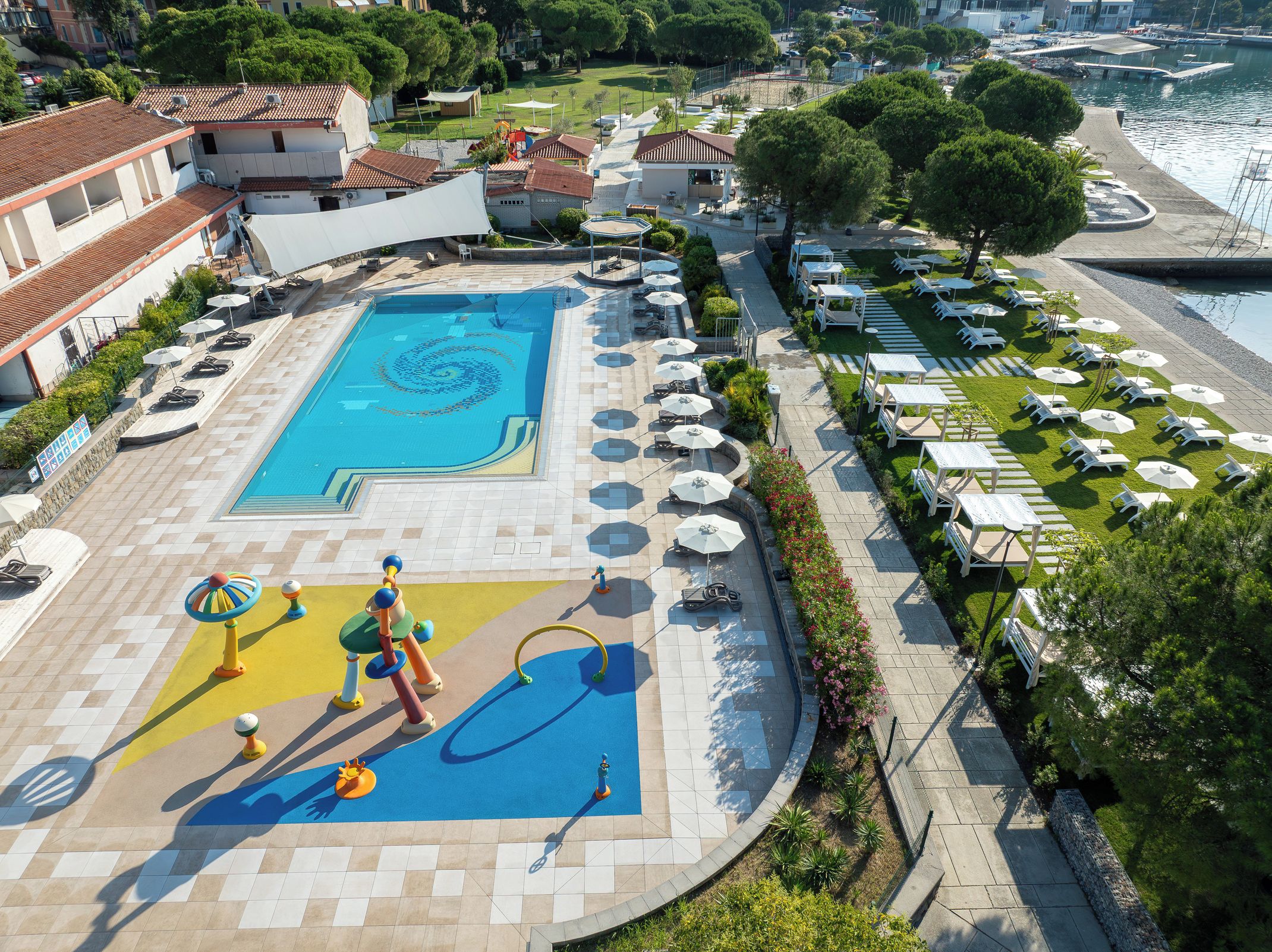 Ein Pool mit spiralförmigem Design, umgeben von Liegestühlen und Sonnenschirmen, mit Blick auf das Meer und Gebäude im Hintergrund.
