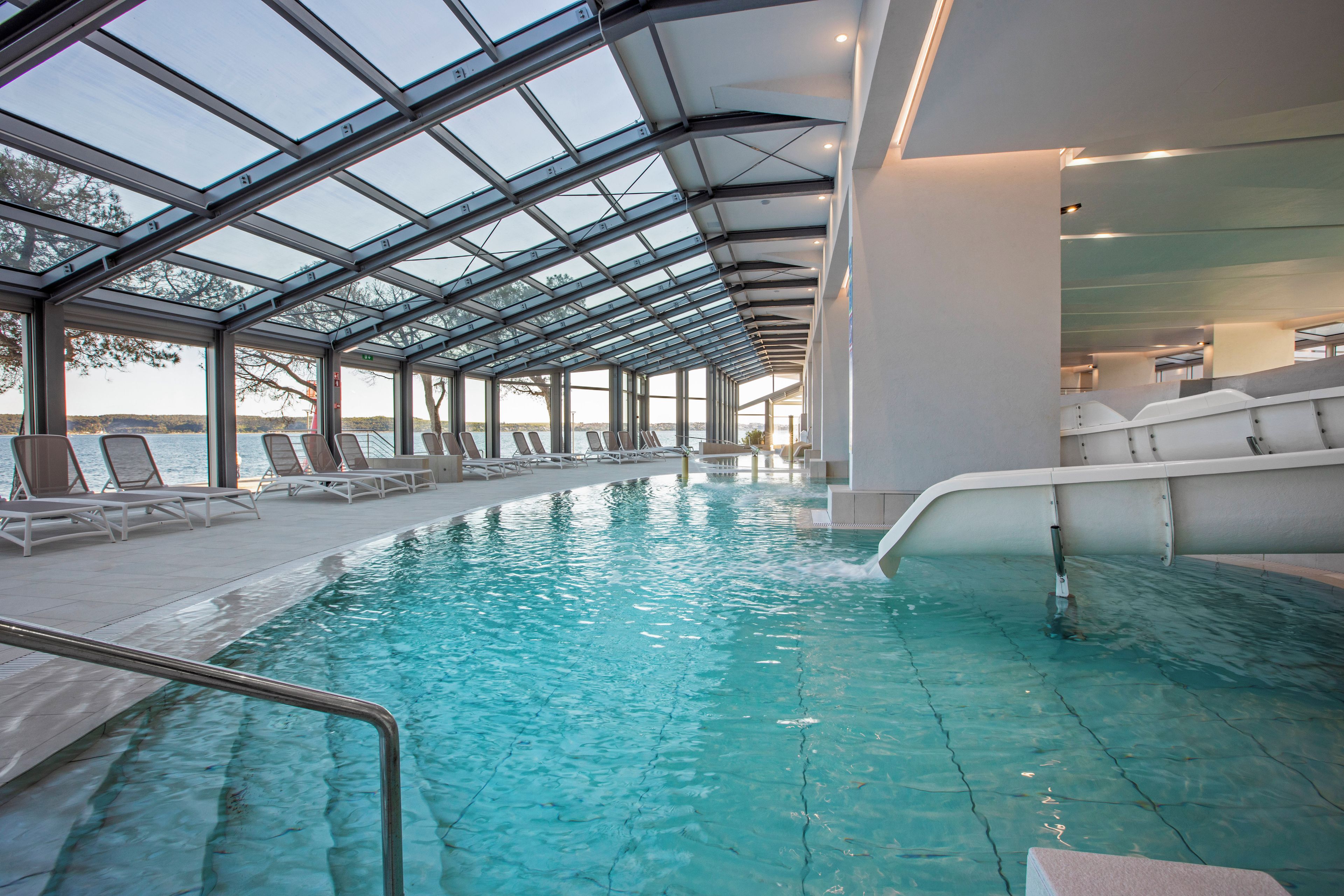 En indendørs swimmingpool med krystalklart vand, omgivet af liggestole og et glastag.