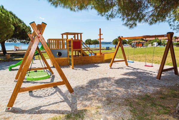 A playground with swings and climbing structures, located near a body of water and trees. - Hotel Vile Park Premium (Piran)