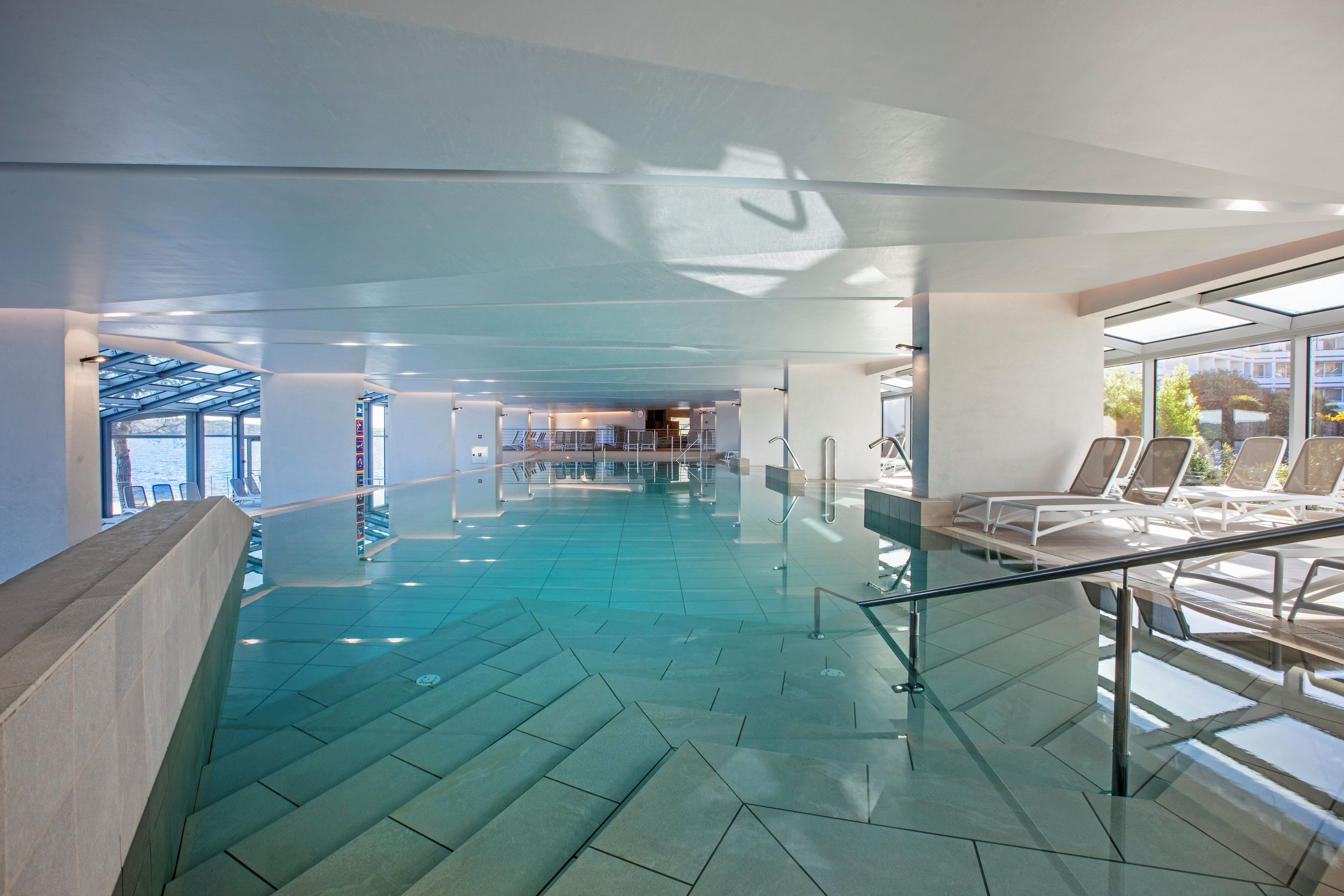 Una piscina interna moderna con pareti in vetro trasparente, un motivo geometrico sul pavimento e un'area relax con poltrone reclinabili.