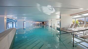 Una piscina interna moderna con pareti in vetro trasparente, un motivo geometrico sul pavimento e un'area relax con poltrone reclinabili.