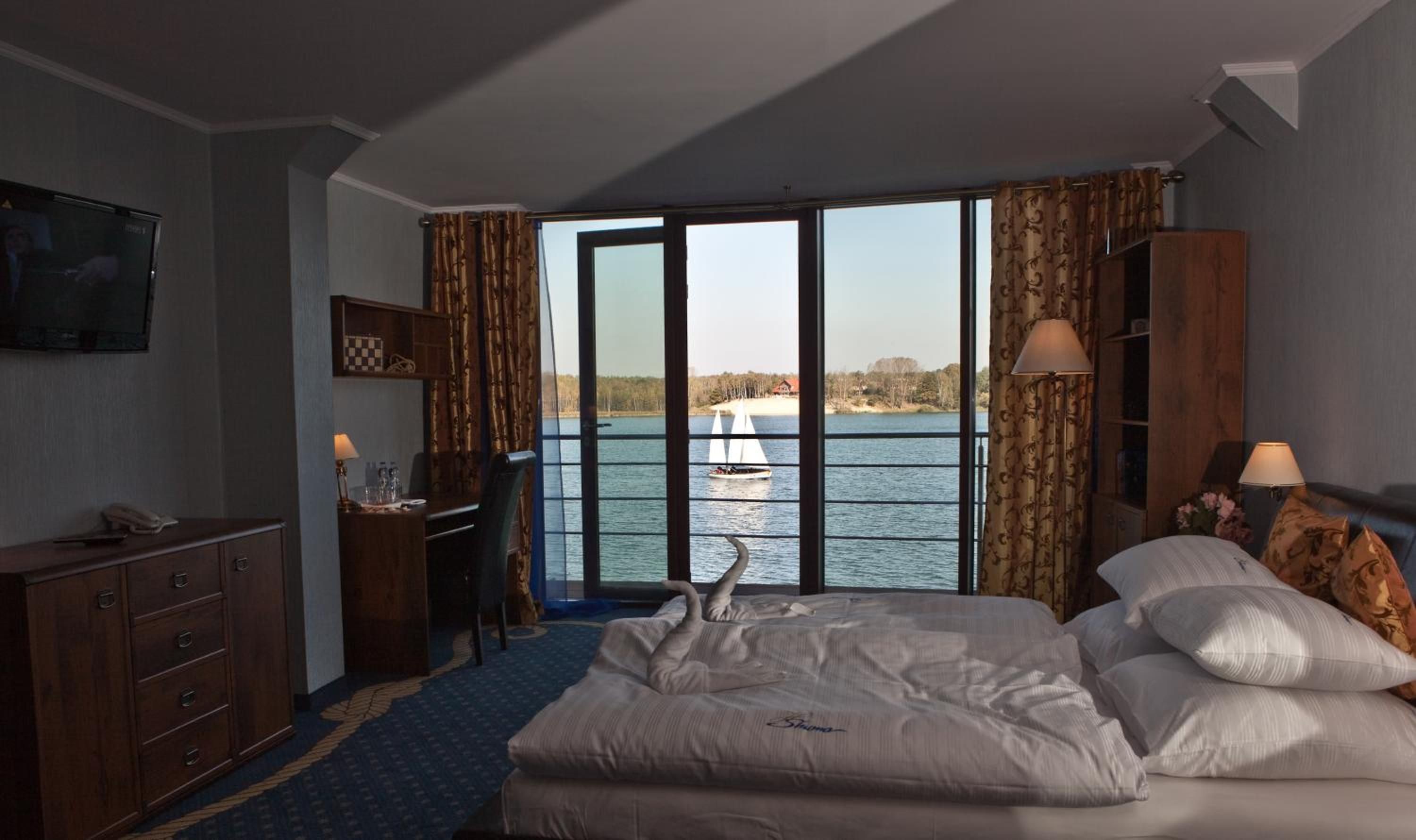 Una habitación de hotel con cama, escritorio, televisión y vistas a un lago con un velero.