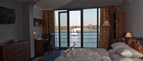 Una habitación de hotel con cama, escritorio, televisión y vistas a un lago con un velero.