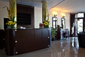 A reception desk with a dark wood finish, flanked by two tall columns with yellow flowers. There are two mirrors above the desk, a potted plant to the right, and a chair in front.