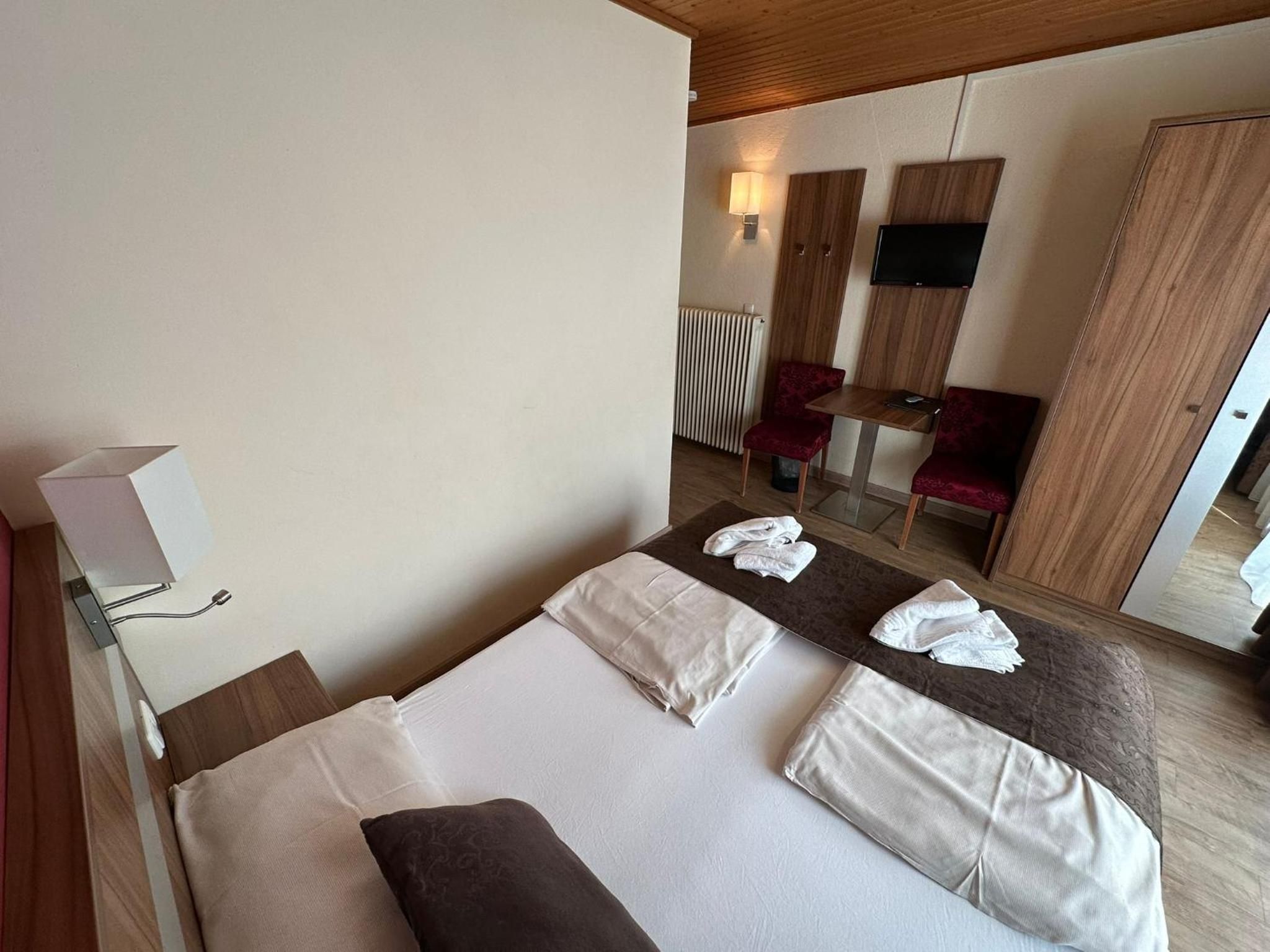 Habitación de hotel con cama, mesita de noche con lámpara, zona de estar con mesa y sillas, y televisor montado en la pared.