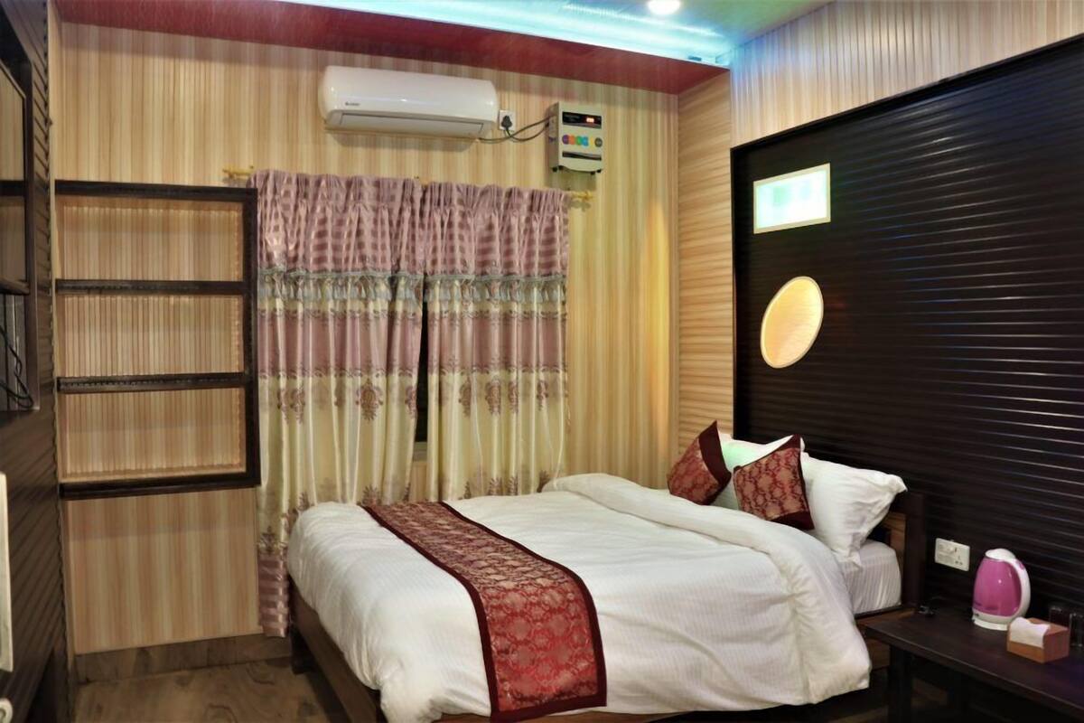 A hotel room with a bed, a bedside table, a lamp, a wall-mounted air conditioner, and a window with curtains.