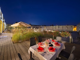 Un espacio al aire libre con mesas dispuestas para una comida, rodeado de plantas y vistas a edificios.
