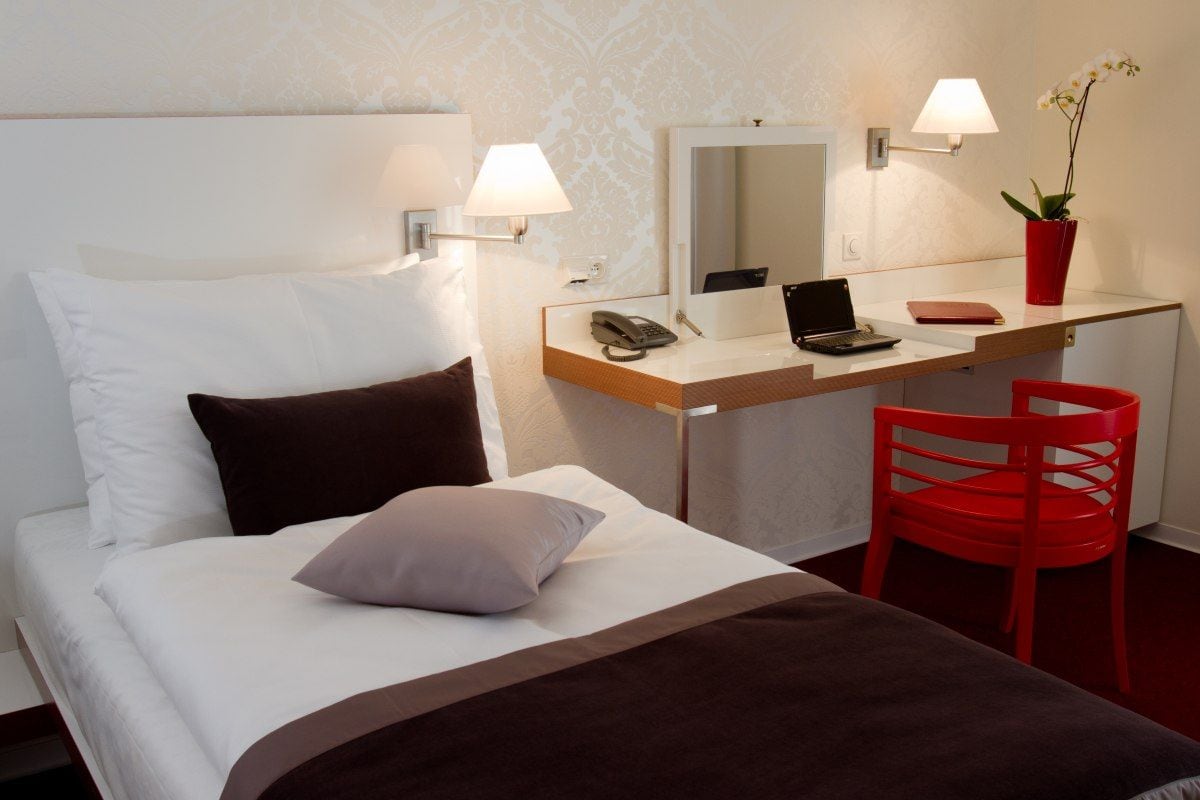 A hotel room with a bed, a desk with a laptop, a phone, and a red chair.