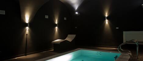 Una piscina interior tenuemente iluminada, con una sola silla de descanso y una escalera de metal.