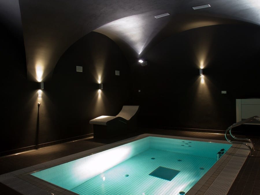 A dimly lit indoor swimming pool with a single lounge chair and a metal pool ladder.