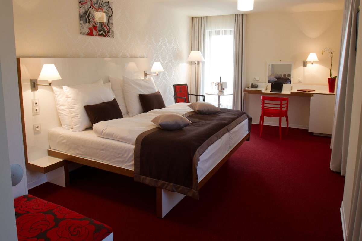 A hotel room with a bed, desk, chair, and a painting on the wall.