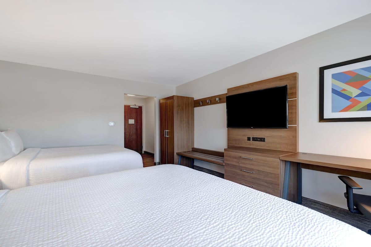 A hotel room with two beds, a wooden desk, a flat-screen TV, and a framed abstract painting on the wall.
