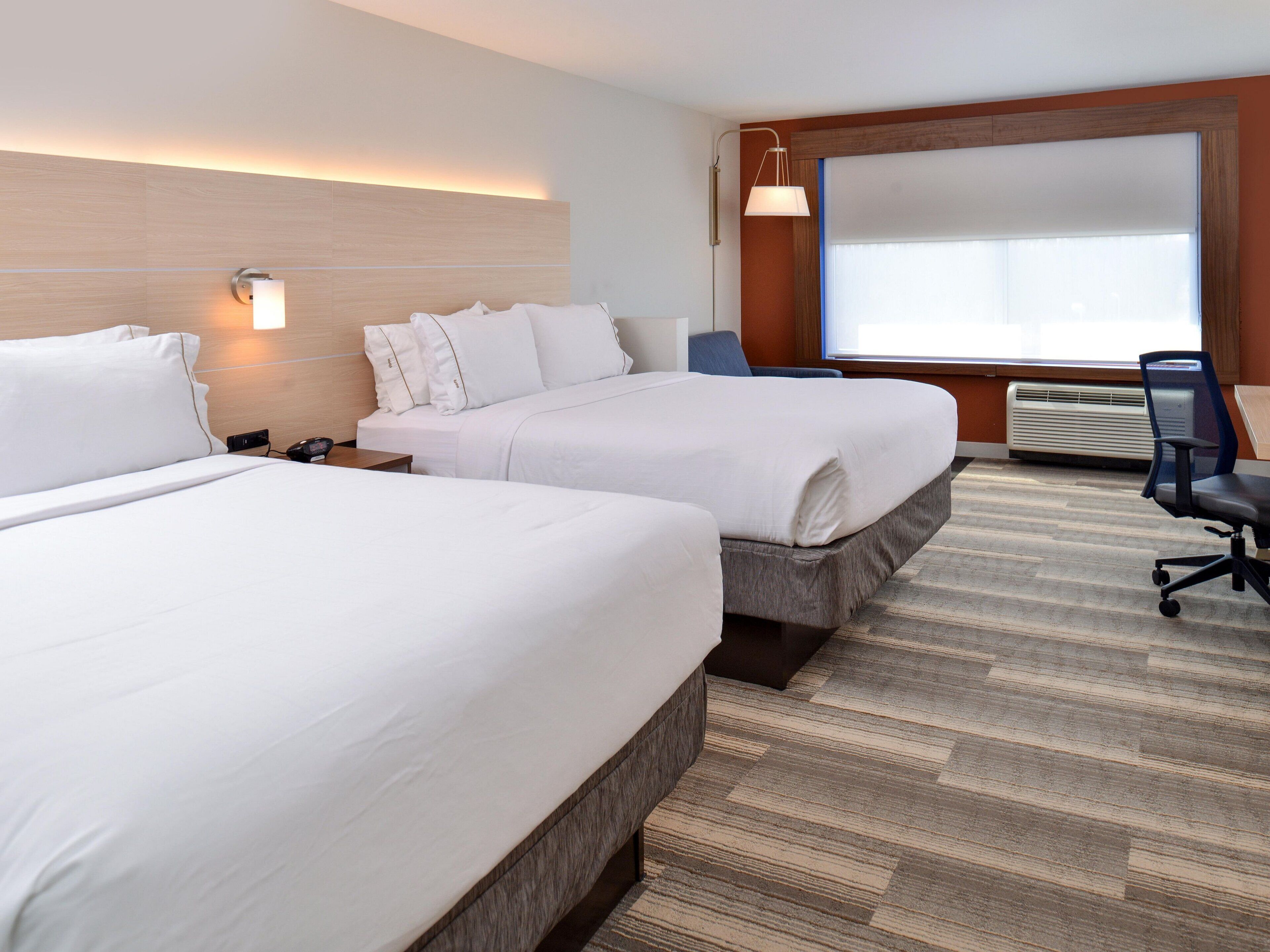 A hotel room with two beds, a desk, a chair, a window with blinds, and a wall-mounted lamp.