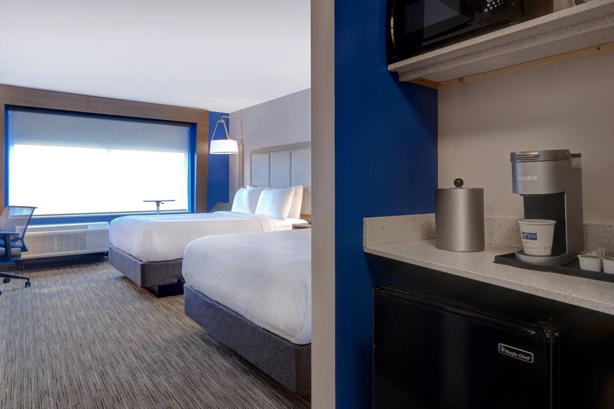 A hotel room with two beds, a desk, a kitchenette with a coffee maker, and a large window.