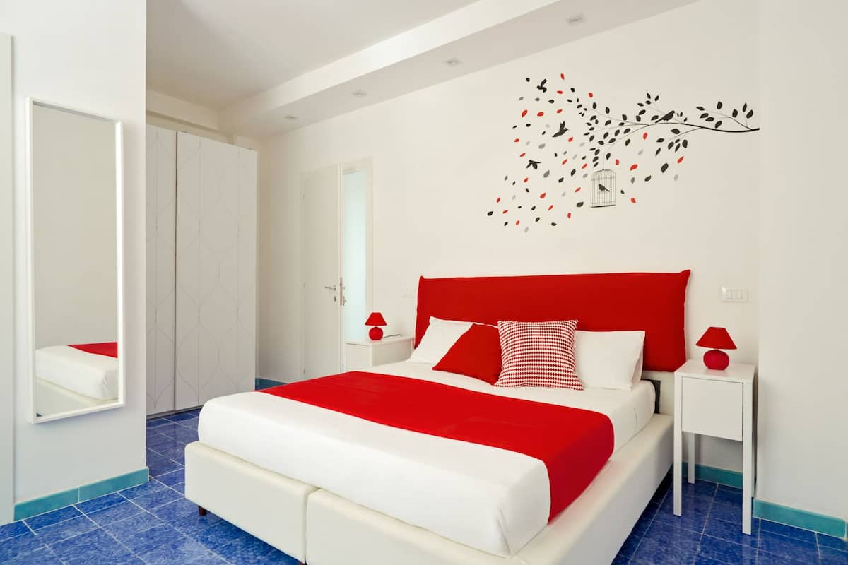 Een moderne slaapkamer met een groot bed, witte muren, een rode accentmuur en een decoratief kunstwerk aan de wand.