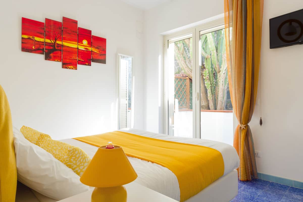 Een hotelkamer met een bed, een gele lamp, een schilderij van een rode zonsondergang en een raam met gordijnen.