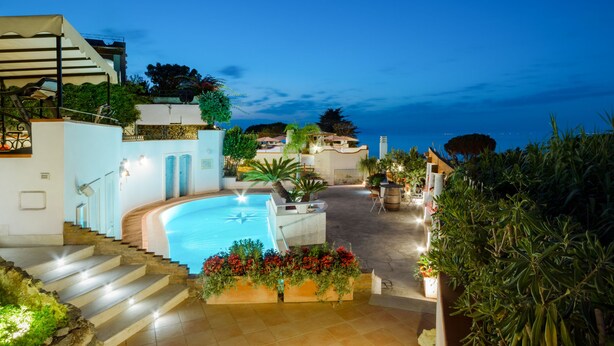 A pool area with steps leading down to it, surrounded by greenery and a view of the sea.