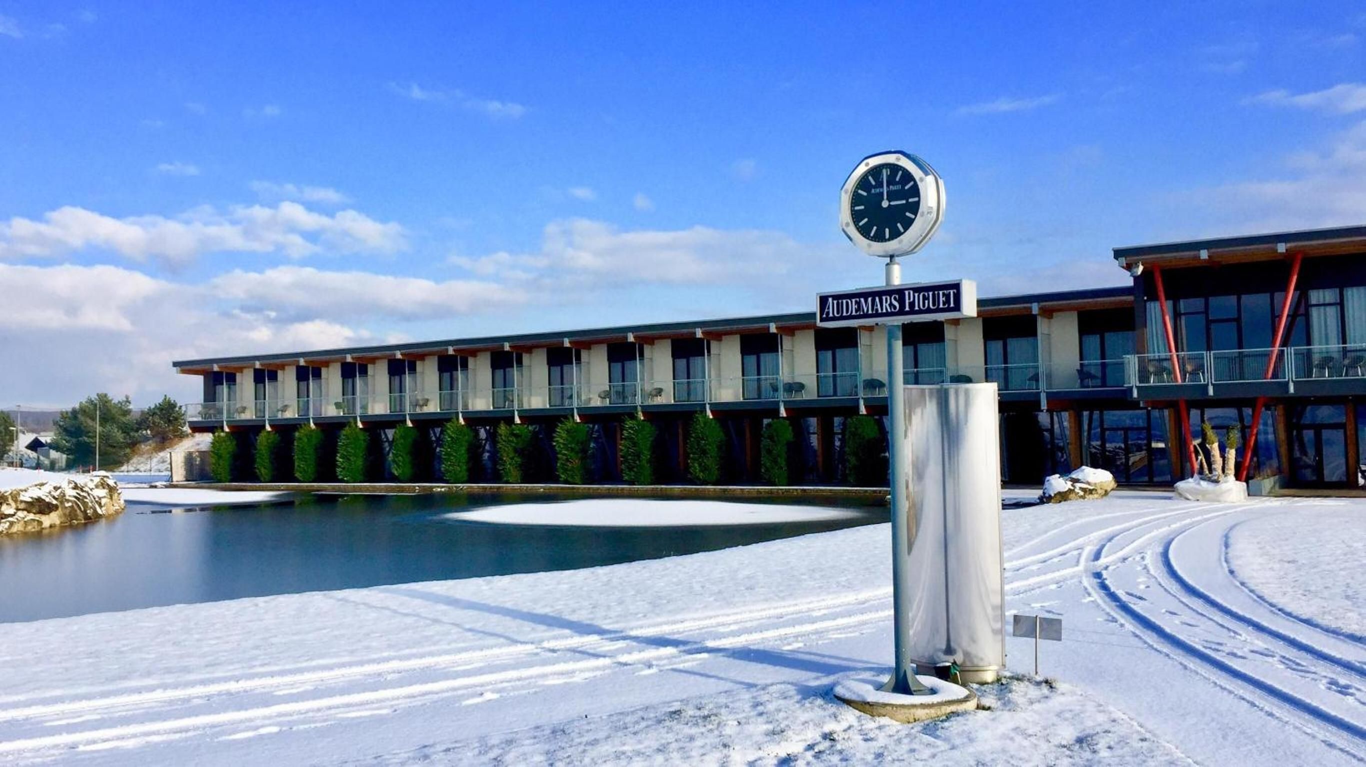 A modern hotel with a clock, a sign reading 'Audemars Piguet,' and a pool in the foreground.