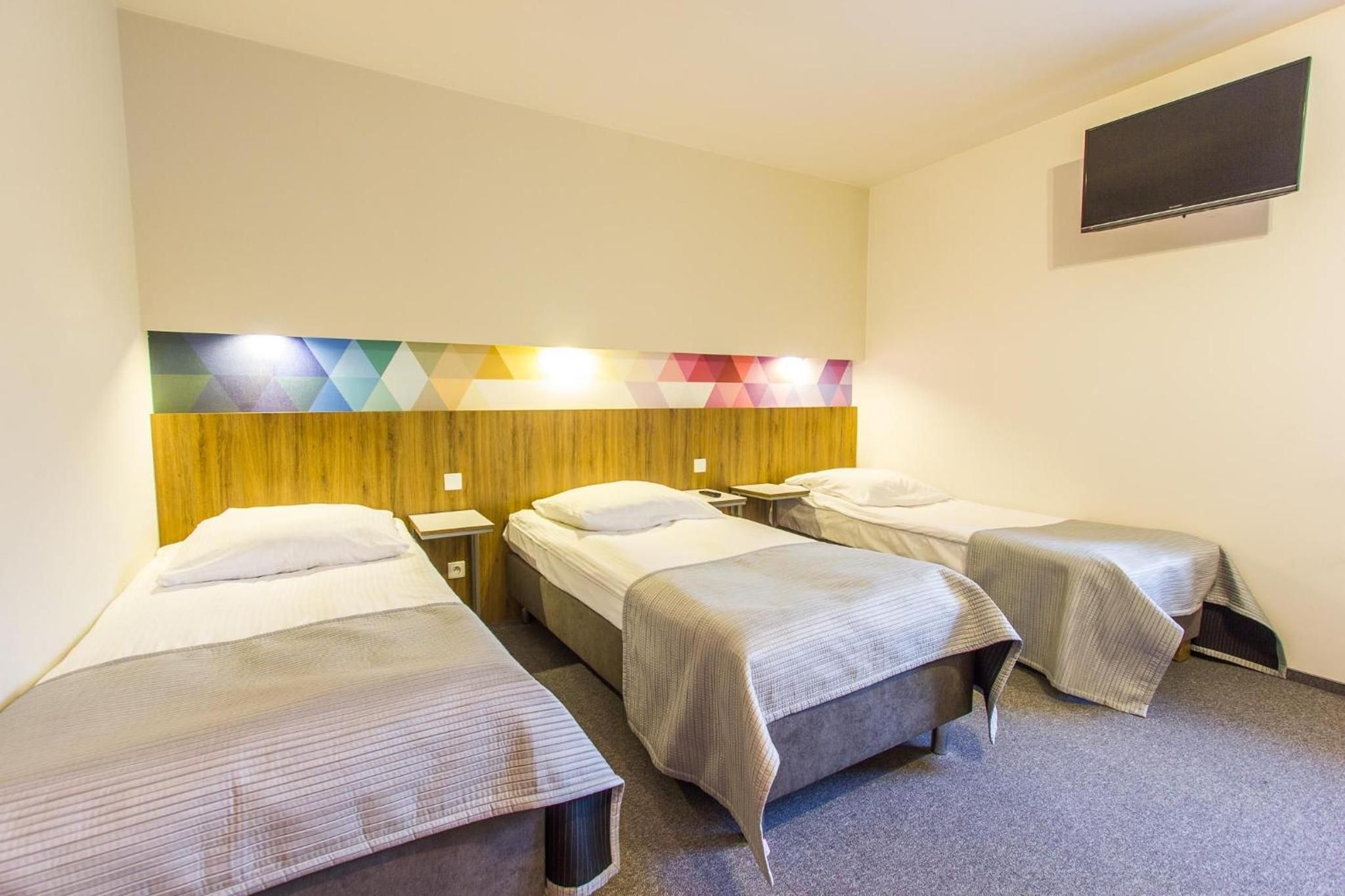 Ein Hotelzimmer mit drei Einzelbetten, einem hölzernen Kopfteil, einem an der Wand befestigten Flachbildfernseher und einer geometrisch gemusterten Leuchte über den Betten.